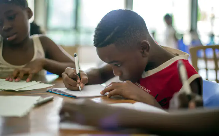 A boy writing on printer paper near a girl